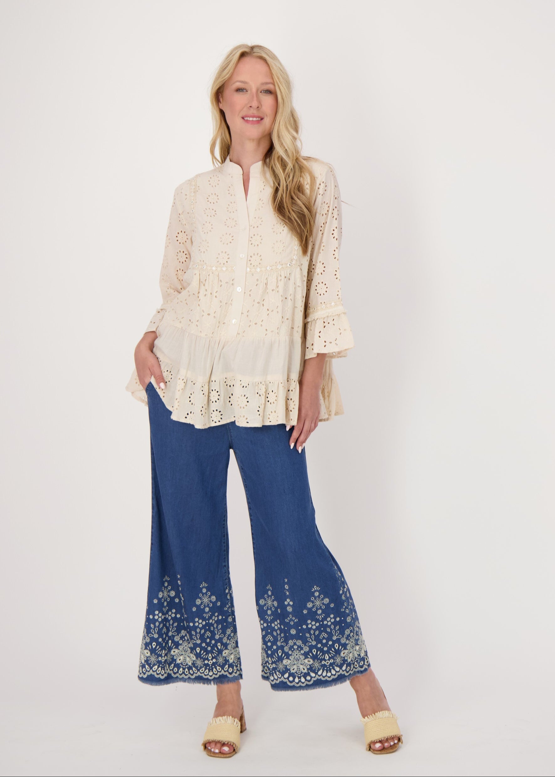 Woman wearing a cream lace top and blue pants with white embroidery on a white background