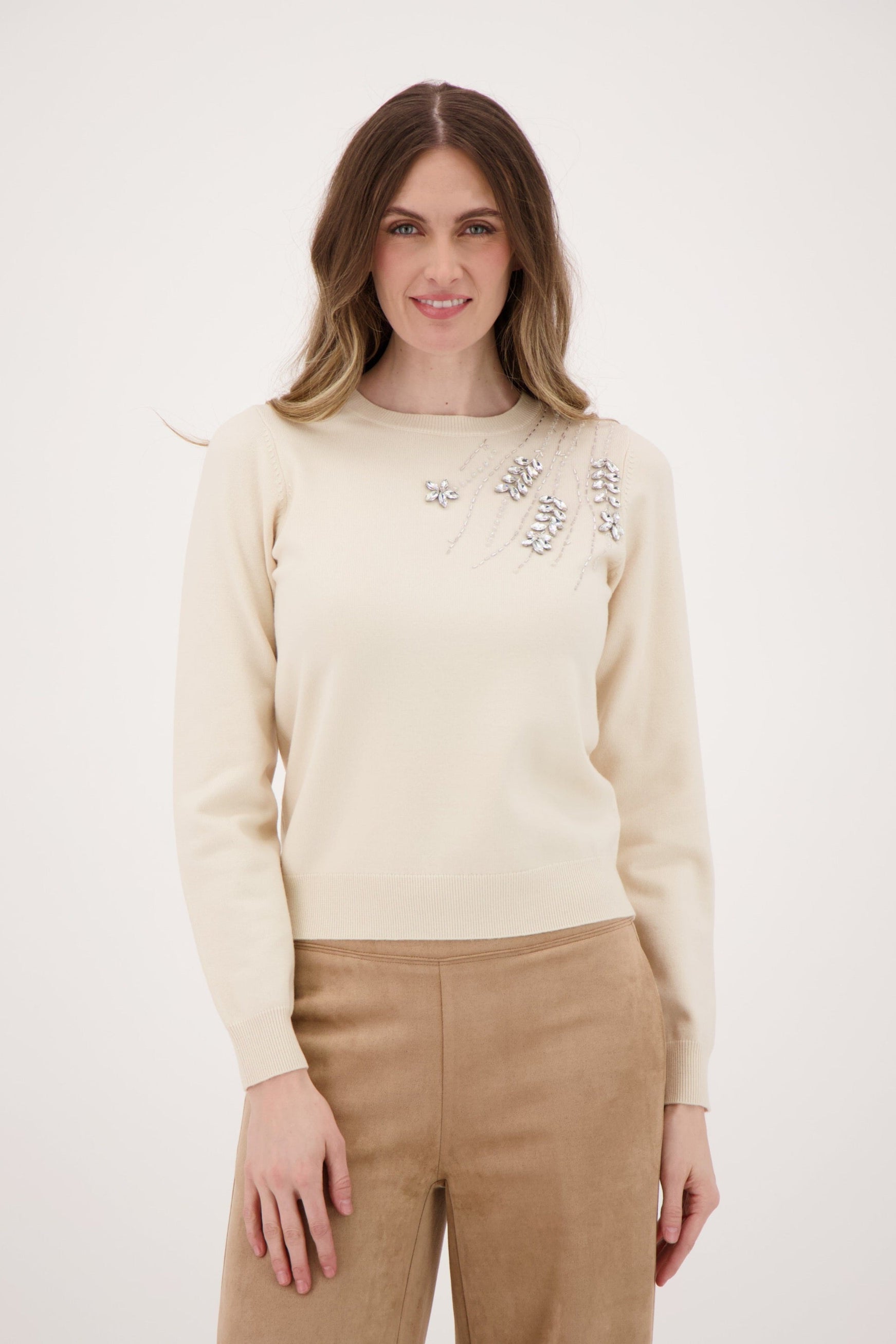 Woman wearing a cream sweater with floral embroidery and beige pants on a white background