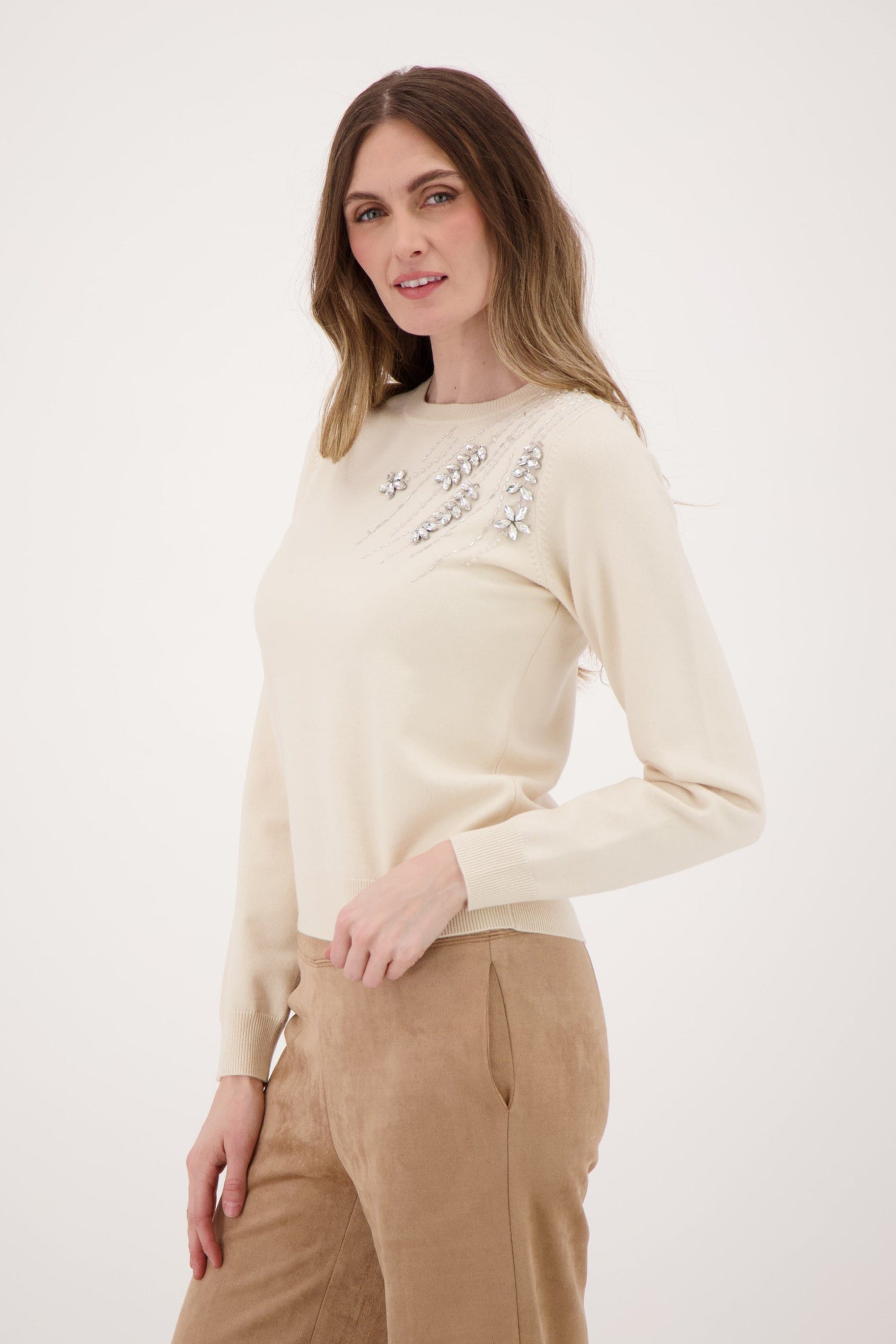 Woman wearing a cream sweater with floral embellishments and beige pants on a white background