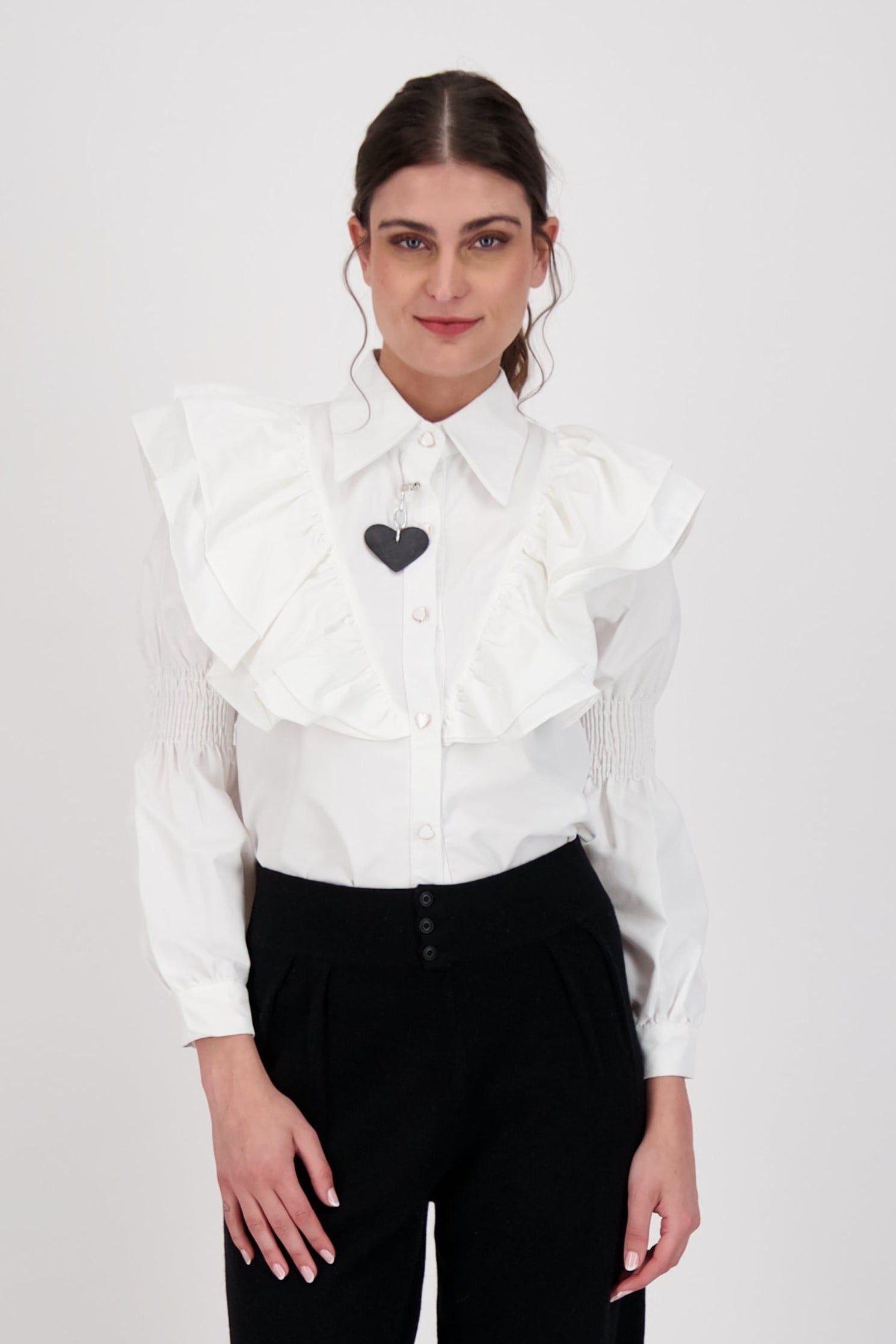 Woman wearing a white blouse with ruffled collar and black heart emblem, paired with black pants on a white background.