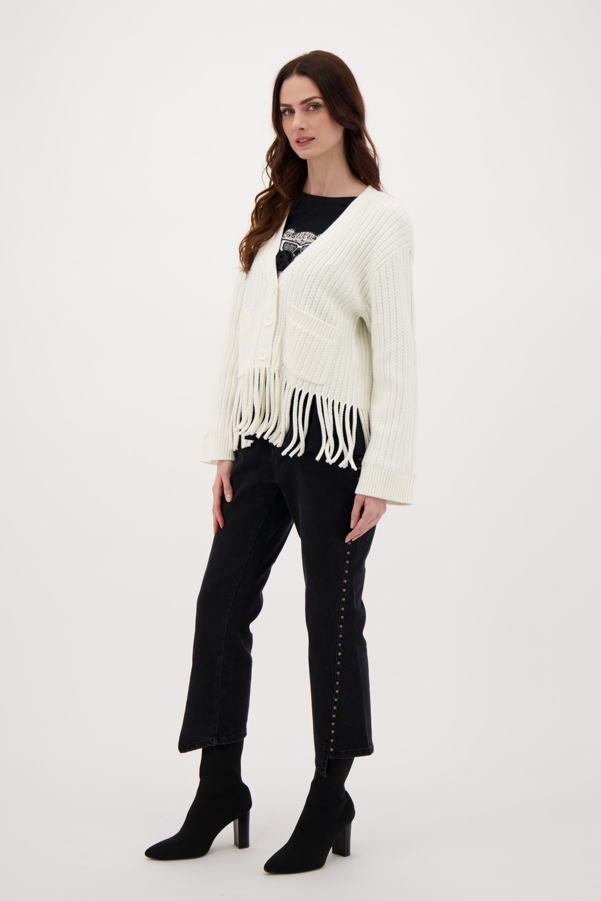 Woman wearing a white cardigan with fringe, black top, pants, and boots on a white background